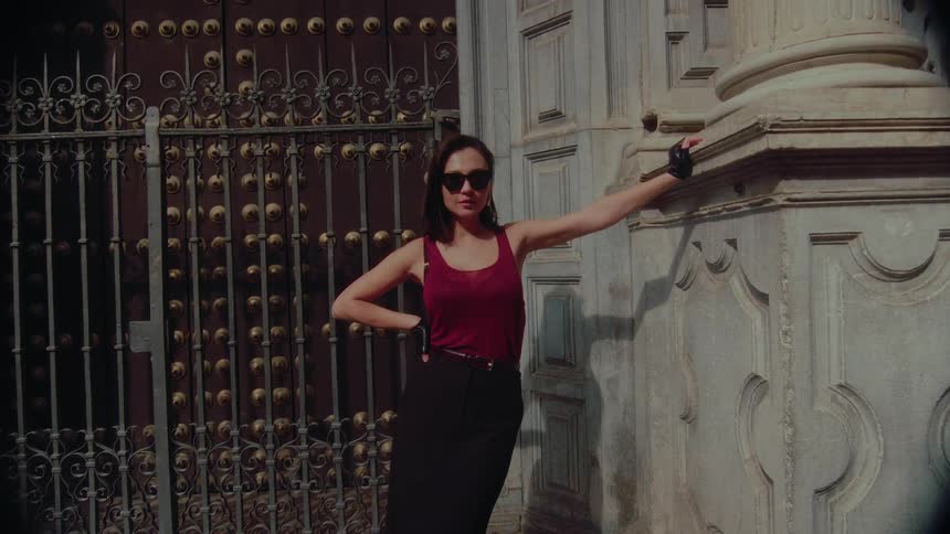 A stylish woman in sunglasses and fingerless gloves confidently poses against an old European building