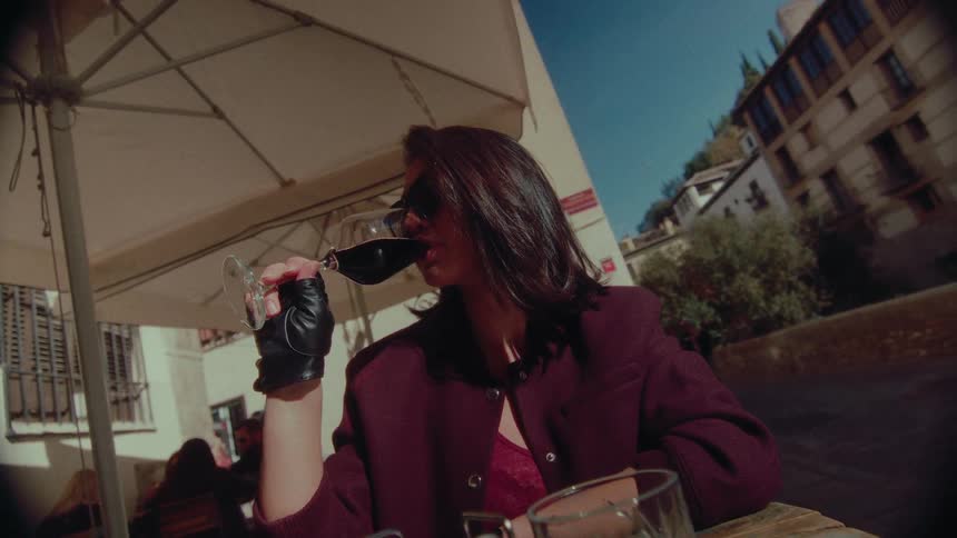 A chic woman in sunglasses slowly sips red wine while relaxing at a sunny outdoor cafe table