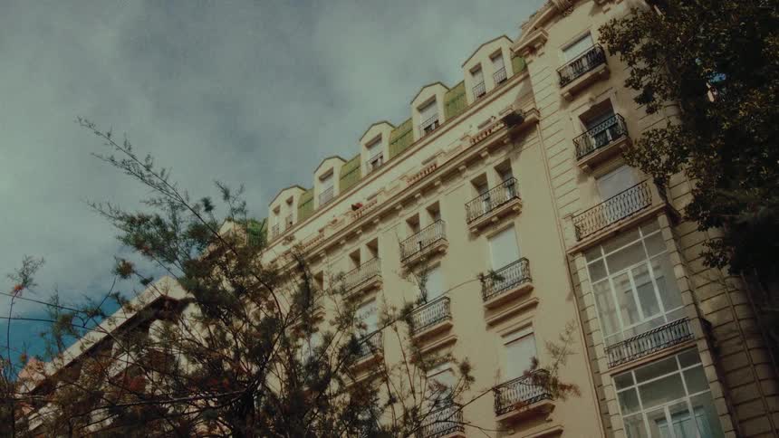 A slow zoom out from a low angle reveals an ornate European apartment building on a cloudy day, framed by trees