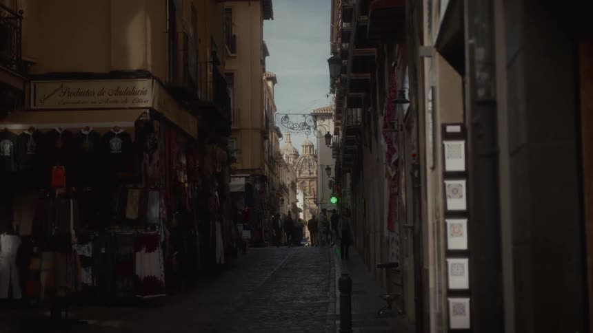 A view down a shaded, narrow cobblestone street in Andalusia, Spain, lined with souvenir shops and leading to a sunlit historic plaza