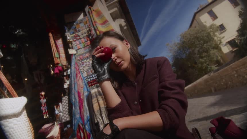 A stylish woman in black leather gloves holds a red apple up to her face while sitting by a colorful European market stall