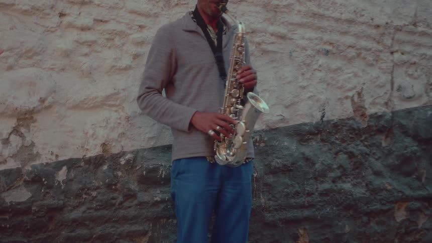 A street musician stands against a weathered stone wall, playing a soulful tune on his saxophone as people pass by