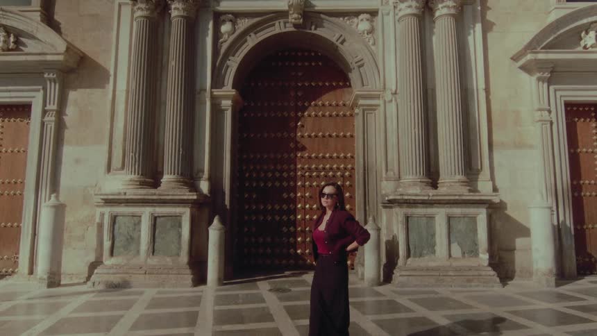 A stylish woman in sunglasses turns and walks toward the grand, studded doors of a historic building in the sunlight