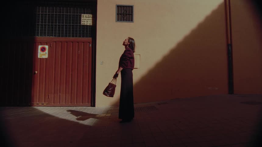 An elegant woman stands alone in a sunlit corner of a quiet urban alley, tilting her head up towards the light