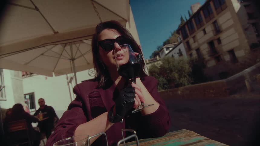 A chic woman in sunglasses slowly sips a glass of red wine at a sunny outdoor cafe in a historic European city