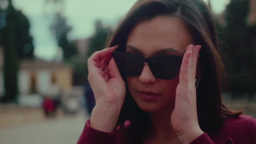A pensive and stylish woman pulls her sunglasses from her purse and puts them on in a public park in slow motion