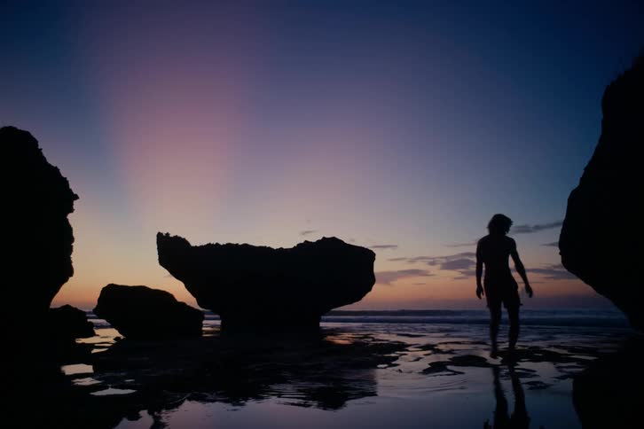 A serene silhouette of a woman walks through shallow water on a rocky beach as the sun sets