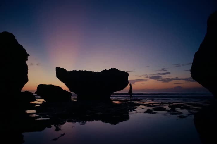 A person's silhouette stands on a tropical beach at sunset, framed by large rocks and calm water