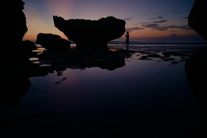 A lone silhouetted figure stands on a rocky beach next to a large rock formation, watching a beautiful sunset with reflections in the tide pools