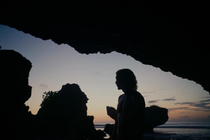 A silhouetted person with long hair stands contemplatively inside a beach cave, looking out at the ocean during a serene sunset