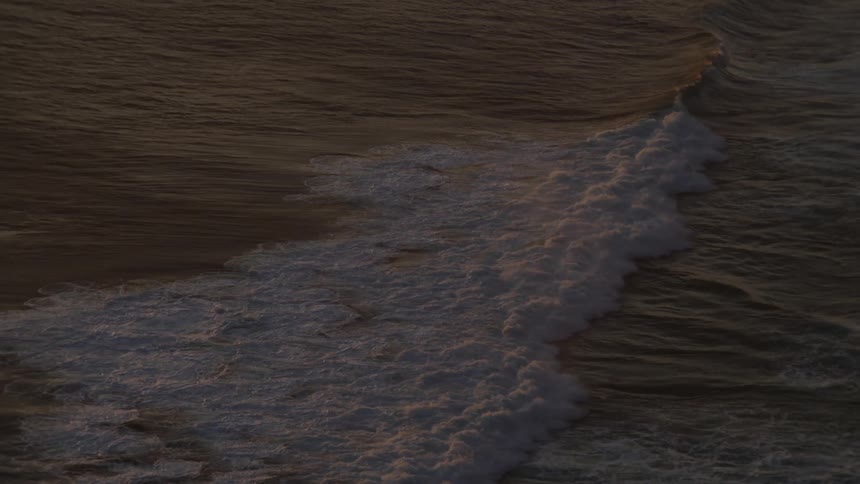 A serene slow-motion high-angle view of a breaking ocean wave illuminated by the soft light of sunset