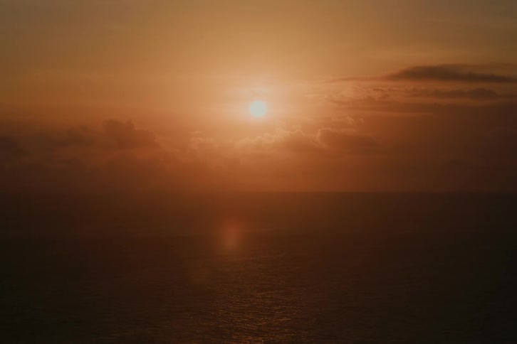 The warm sun sets over a calm, hazy ocean, reflecting its golden light on the water's surface