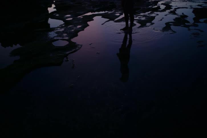 A person's silhouette is reflected in the dark, still water of a tide pool at twilight
