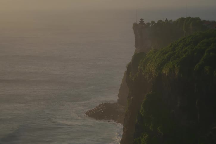 A majestic temple sits atop a lush green cliff as gentle ocean waves roll in during a hazy golden sunset
