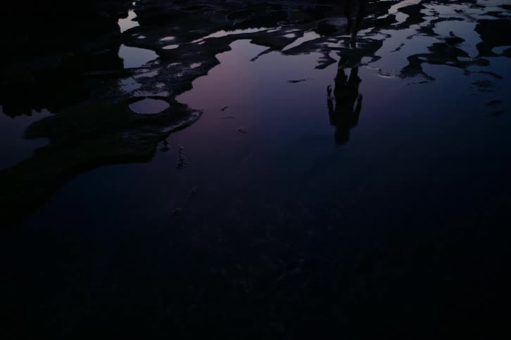 The mysterious reflection of a person appears in a dark, still tide pool as they walk into the water at twilight