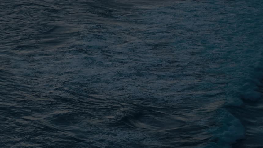 A single wave crests and rolls across the dark, moody ocean surface in an abstract slow-motion shot