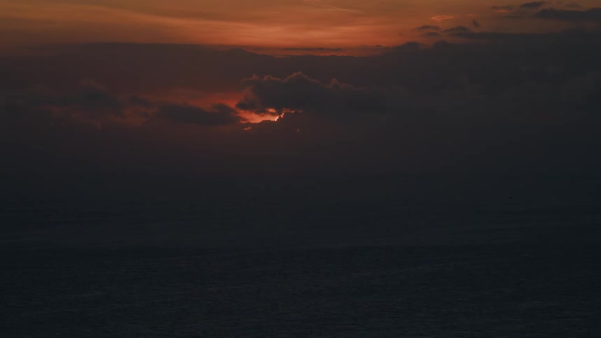 The last light of a red sunset breaks through a dark, cloudy sky over the vast, dark ocean