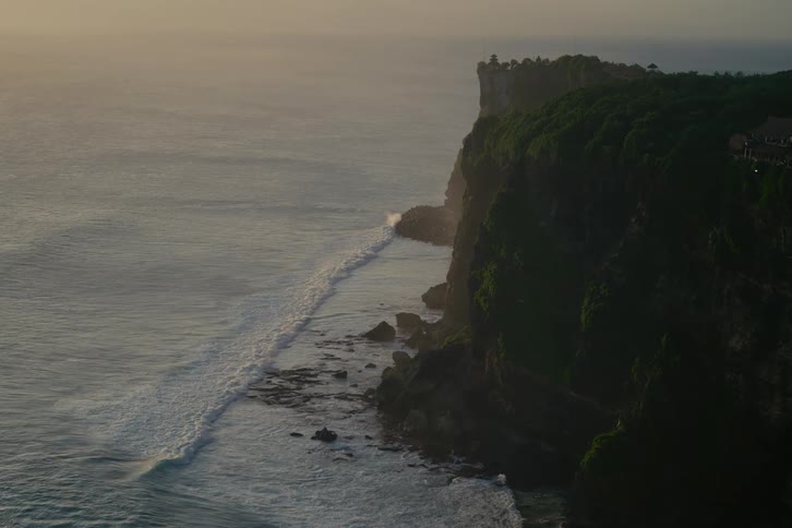 Gentle ocean waves wash against a lush green cliffside during a tranquil golden hour sunset