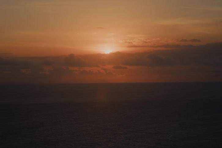 A serene wide shot captures the golden sun setting over the calm ocean horizon