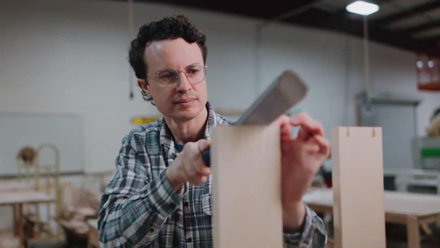 A focused male carpenter with glasses carefully uses a hand saw to cut a precise joint in wood inside a busy workshop