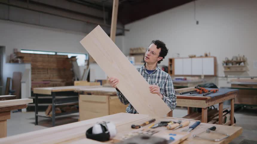 A skilled carpenter carefully inspects a long wooden plank for quality inside his busy workshop