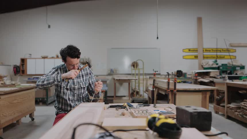 A skilled woodworker carefully carves wood with a chisel and hammer in slow motion within his workshop