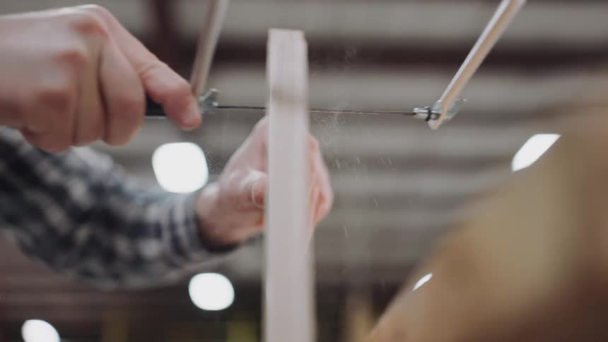 A close-up slow-motion shot captures a woodworker's hands meticulously cutting a piece of wood with a coping saw in a workshop
