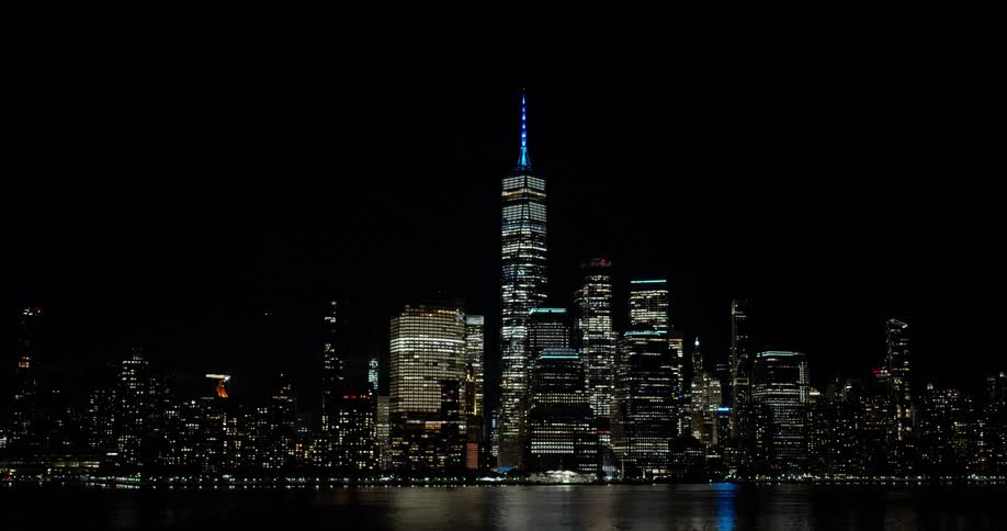 The iconic Lower Manhattan skyline, featuring the illuminated One World Trade Center, glows against the dark night sky with its lights reflecting on the water
