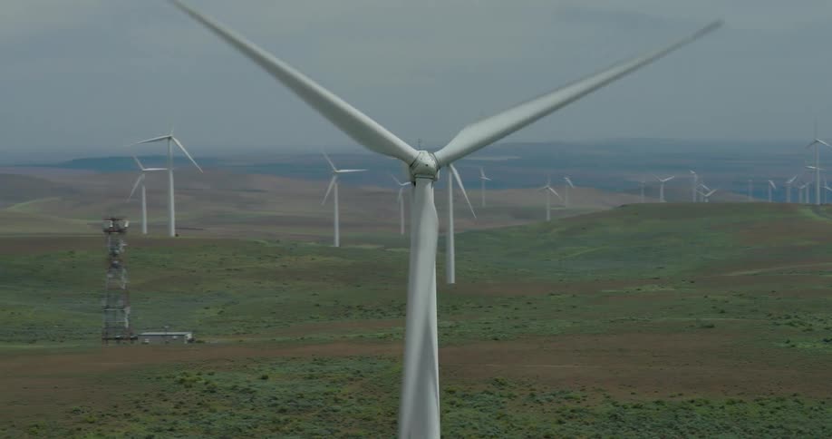 An aerial shot circles a massive wind turbine as it spins, with a large wind farm visible across the vast, rolling landscape