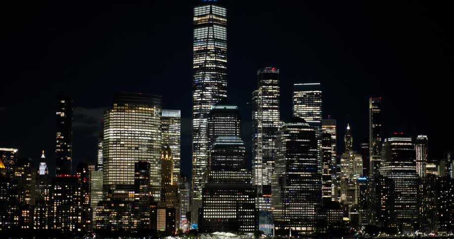 A stunning nighttime aerial view zooms out from the One World Trade Center to reveal the entire illuminated Lower Manhattan skyline and its reflection on the water