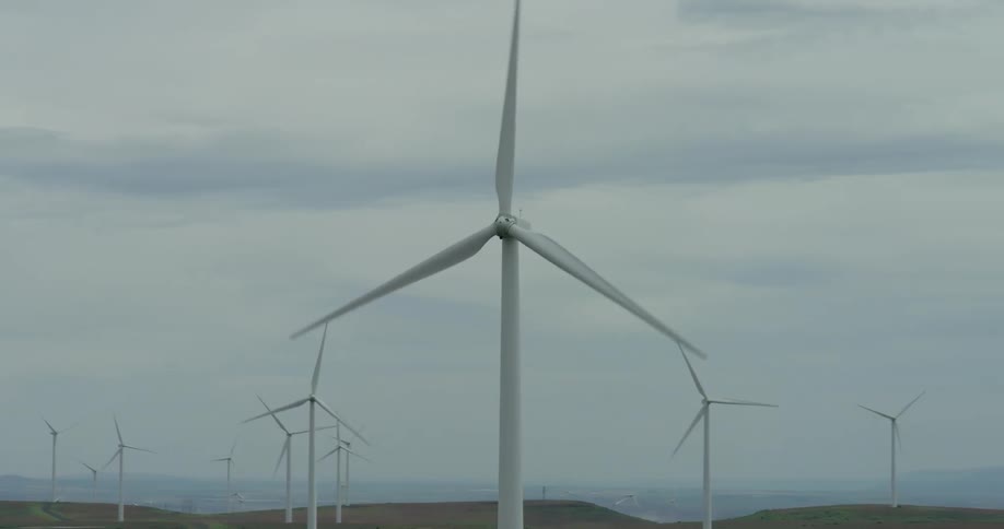 A close-up view of a large wind turbine spinning slowly pulls back to reveal a vast wind farm on rolling hills under an overcast sky
