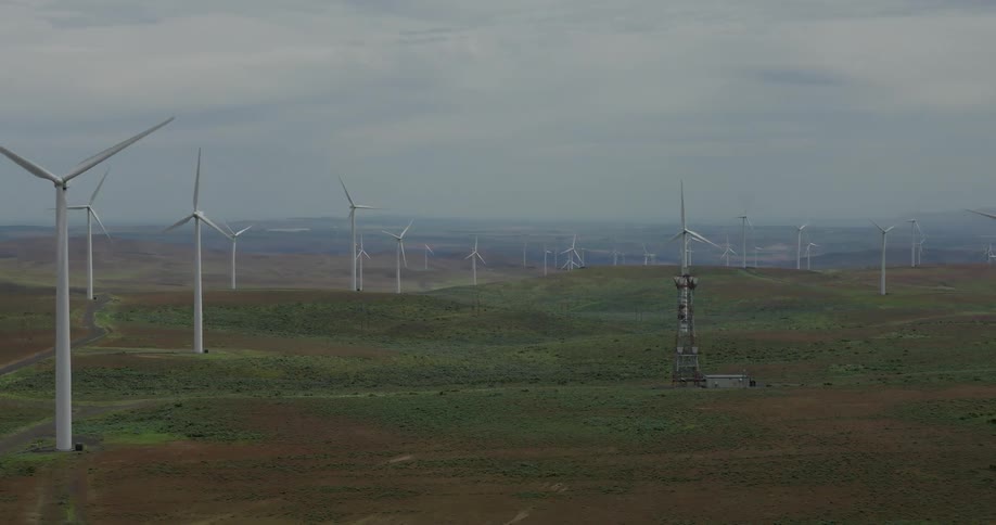 A slow aerial dolly shot reveals a vast wind farm with turbines spinning across rolling green hills under an overcast sky