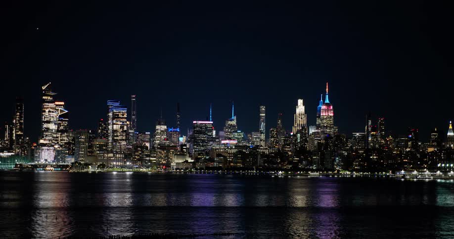 The iconic New York City skyline glitters at night as the camera slowly zooms out, revealing the majestic cityscape and its reflection on the water