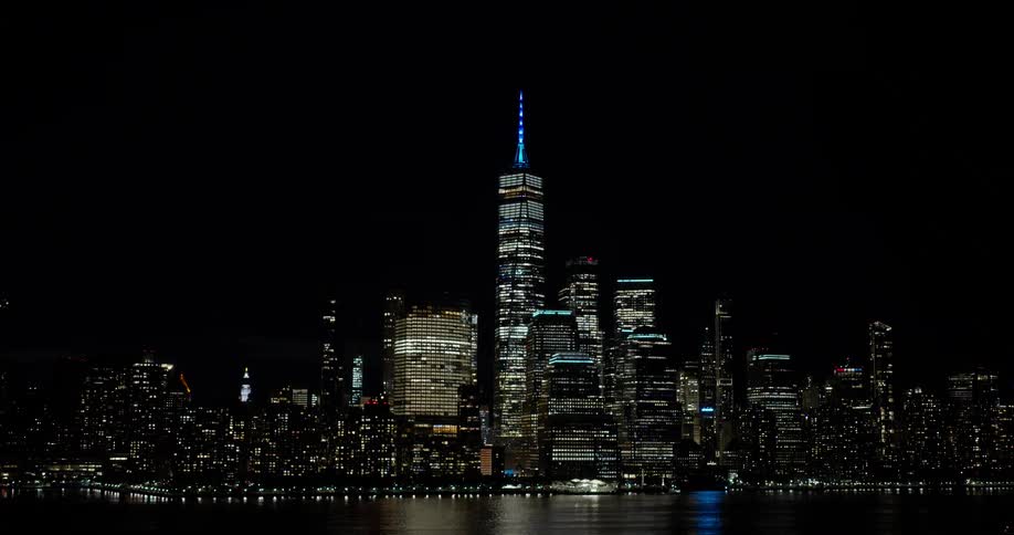An iconic wide shot of the illuminated New York City skyline at night slowly zooms out over the water