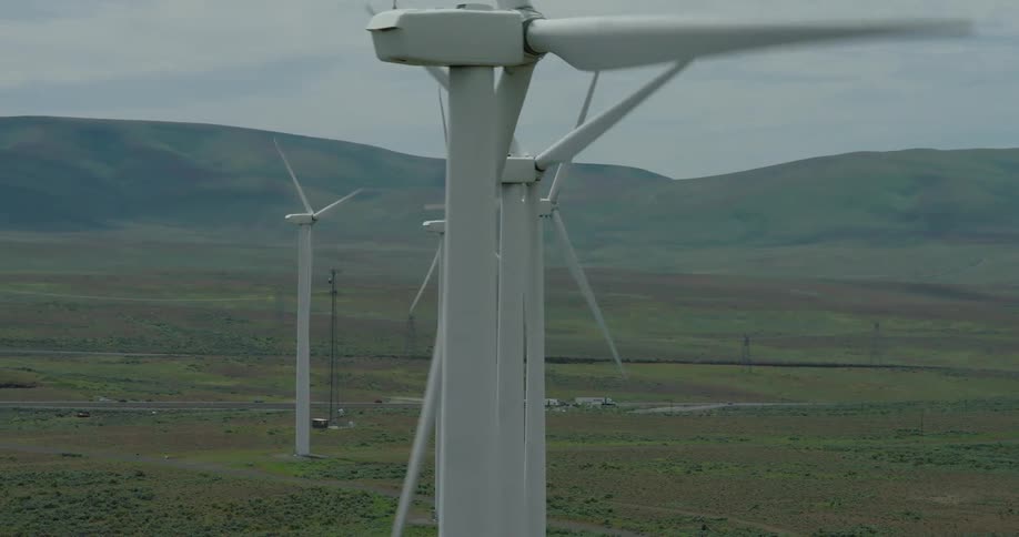 An aerial shot slowly pans across a row of large wind turbines spinning and generating power on a rural wind farm