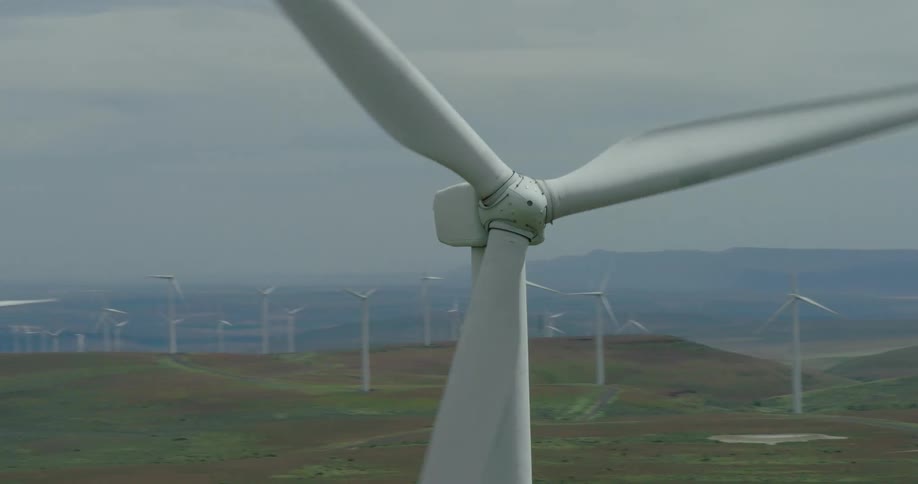 An aerial view pulls back from a close-up of a single spinning wind turbine to reveal a sprawling wind farm across rolling green hills