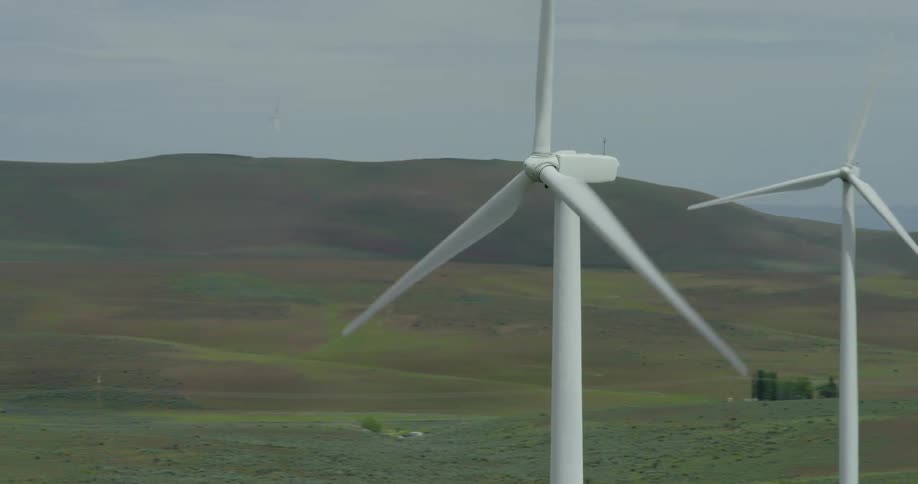 An aerial view orbits a spinning wind turbine in a vast wind farm set amongst rolling green hills on an overcast day