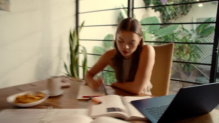 A focused young woman studies at a wooden table with a laptop and textbooks in a brightly lit room