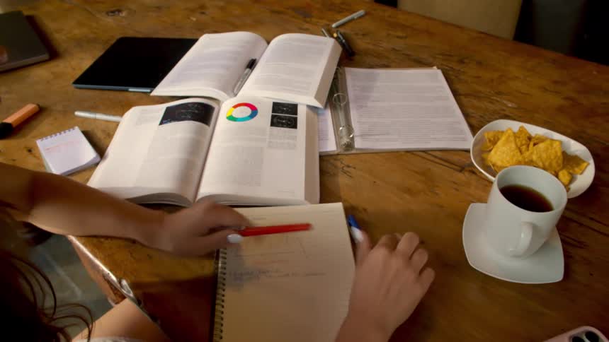 Over-the-shoulder slow motion shot of a young woman studying from a textbook and taking notes at a wooden table