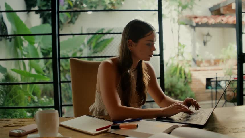 A thoughtful young woman works on her laptop at a desk surrounded by books and overlooking a beautiful garden