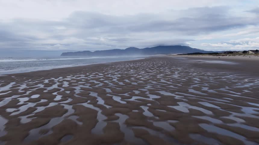 A serene, low aerial flight backward over a vast, wet beach with beautiful sand patterns under a cloudy sky
