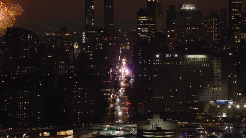 Fireworks explode over a bustling city street at night as traffic moves below the illuminated skyline