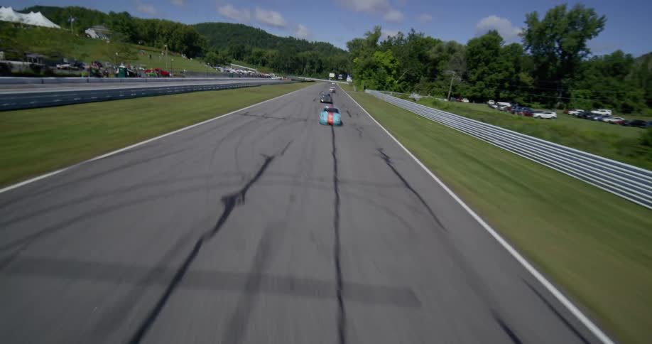 A dynamic low-angle aerial view follows vintage race cars speeding down the main straight at Lime Rock Park on a sunny day