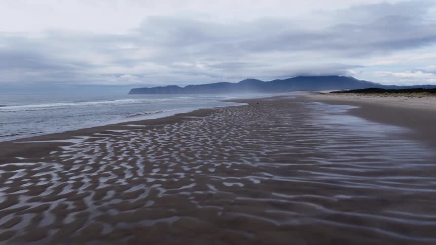 A low aerial flight pulls back over the vast, moody landscape of a beach at low tide, revealing intricate water patterns in the sand