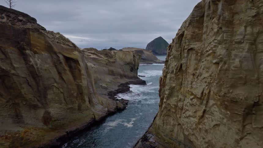 Dramatic aerial footage flying through a rugged sea arch and canyon on a cloudy day along the Oregon coast
