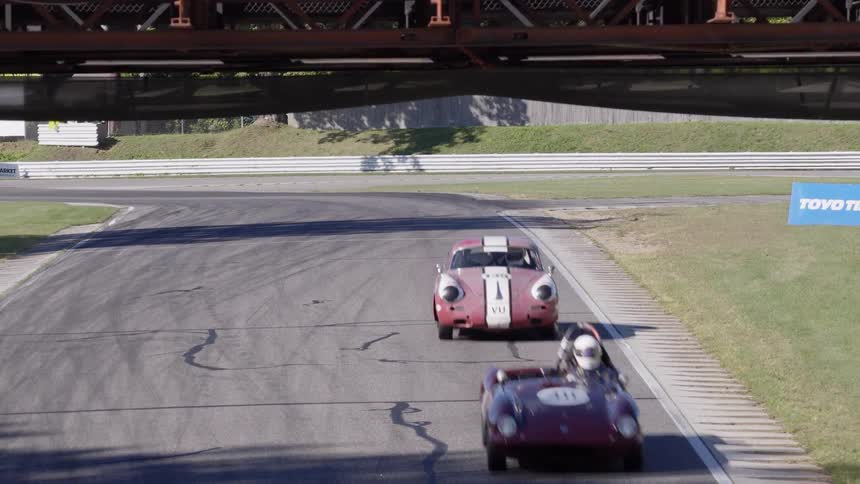 A variety of classic and vintage race cars speed towards the camera and under a bridge on a professional race track