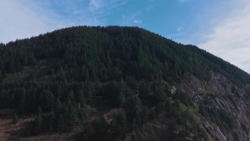 An aerial view shows a white car driving on a winding road carved into the side of a mountain covered in a dense evergreen forest