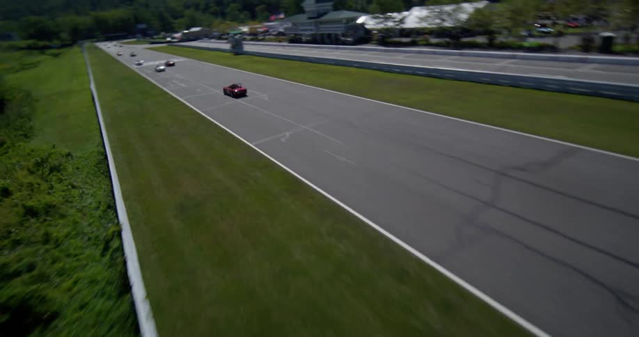 An exhilarating aerial view follows vintage race cars as they speed down the straightaway at Lime Rock Park on a sunny day