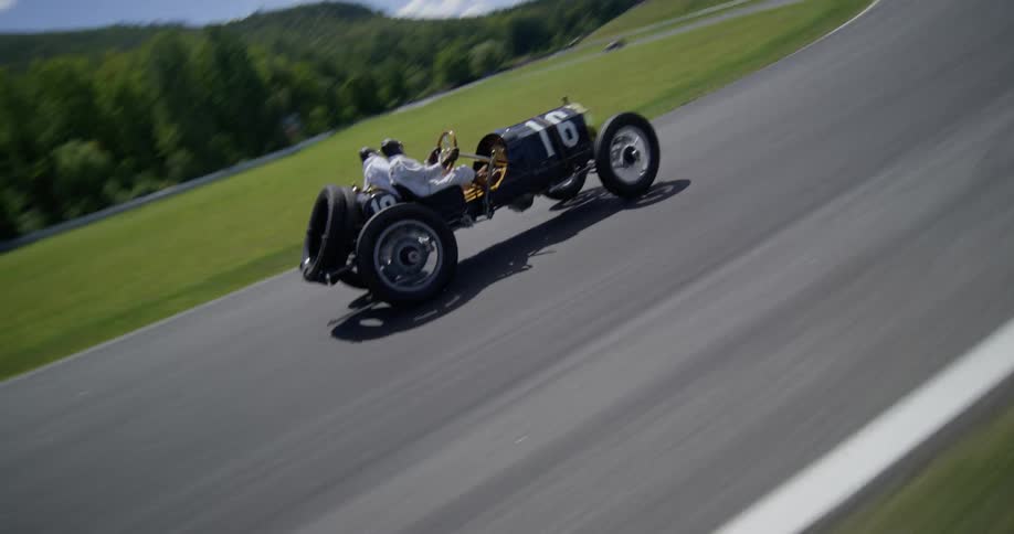 A dynamic aerial shot follows a vintage number 16 race car as it speeds around a turn on a professional racetrack during a classic motorsports event