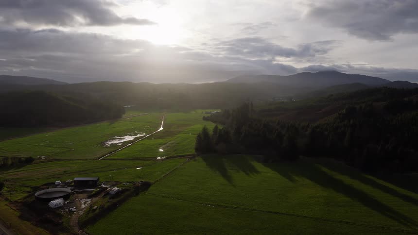 Sunlight bursts through dramatic clouds over a lush green farming valley, casting beautiful light and shadows across the landscape